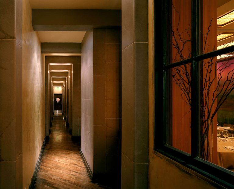 A dimly lit, narrow hallway with tan walls and wooden floors leads to a door with a circular window. To the right, a window reveals a warmly-lit dining area with set tables and decorative branches. Ceiling lights evenly illuminate the corridor.